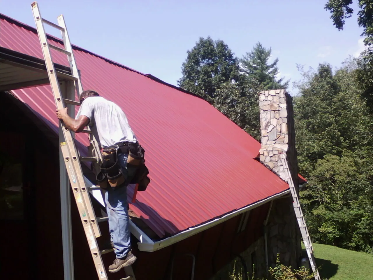 Roofer on a ladder beside a red metal roof and stone chimney — Soffit & Fascia Repair by Roofing Brothers in Apulia Station